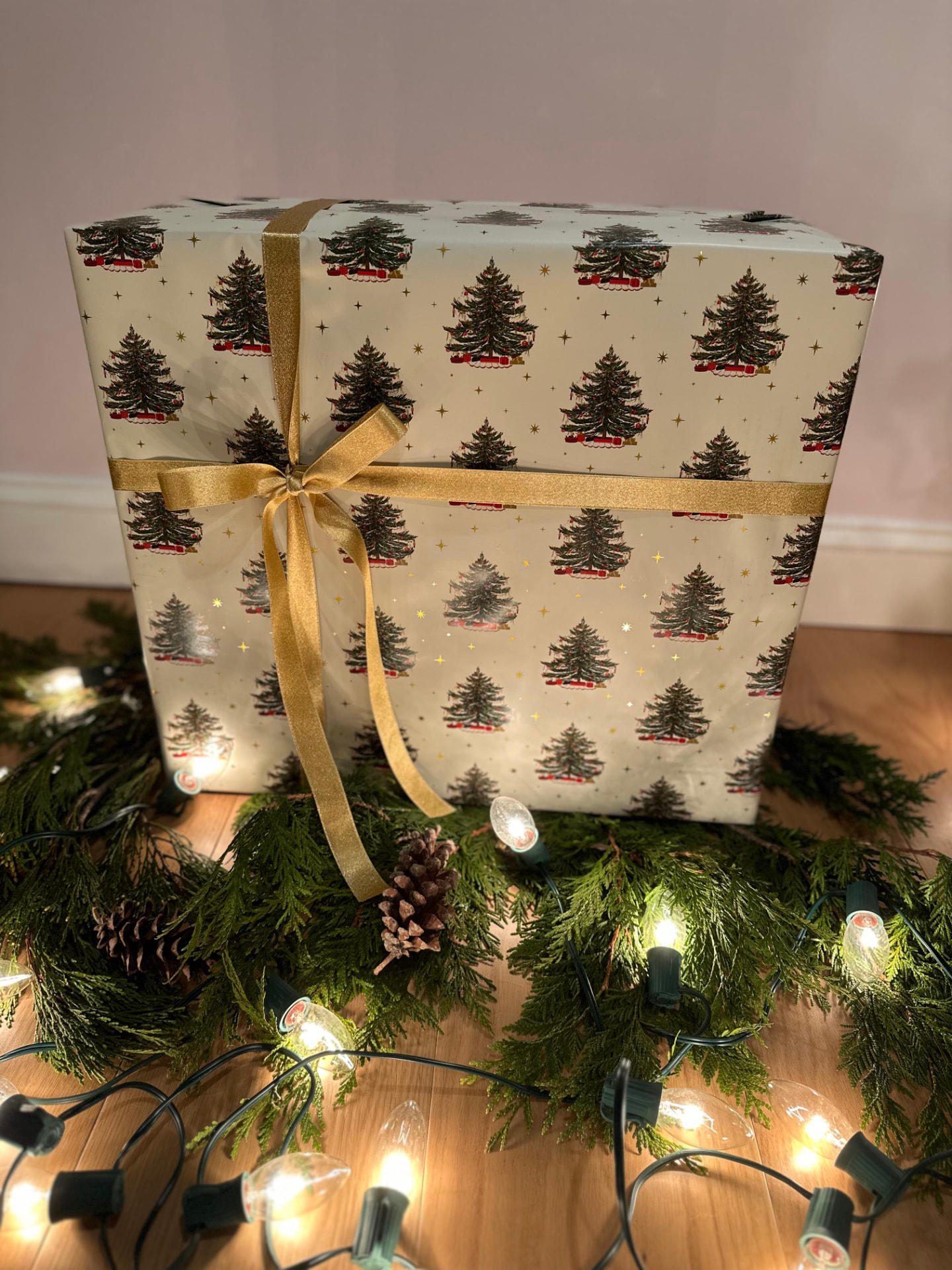 Large holiday gift wrapped in cream Christmas-tree paper with a gold ribbon bow, nestled in garland and lights